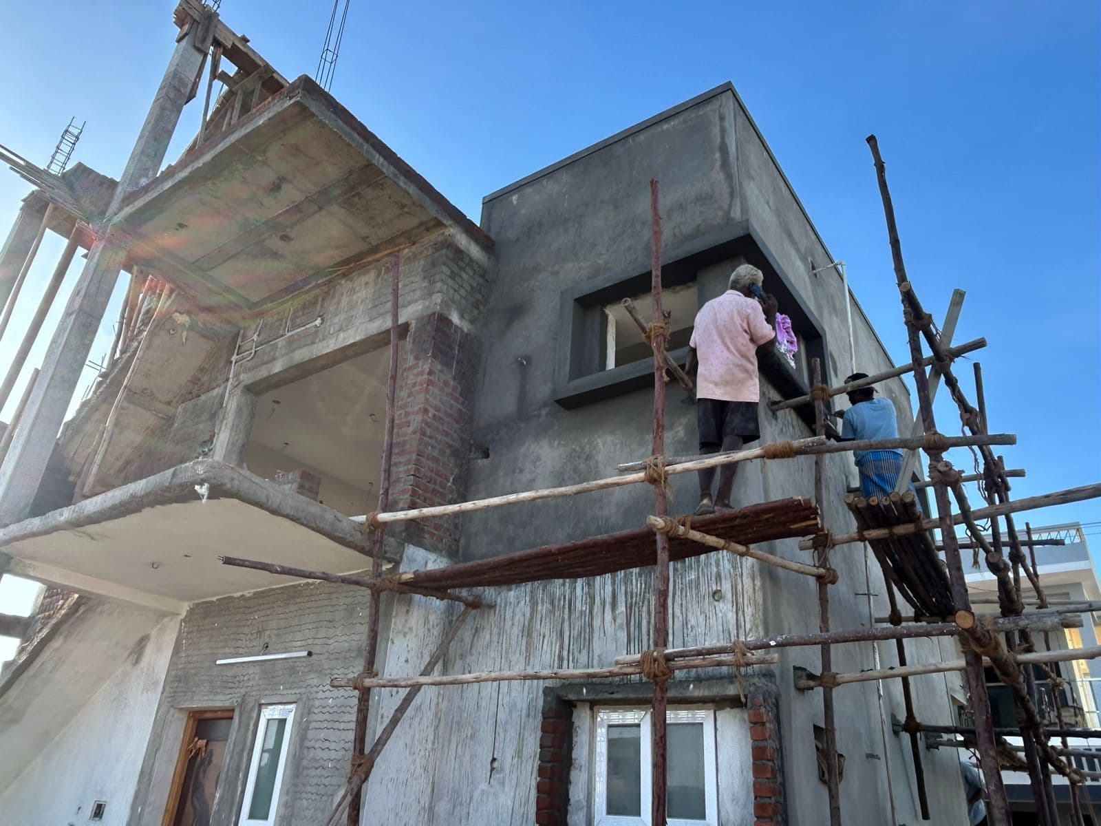 Masons on bamboo scaffolding plastering the external walls of the Deeran Nagar residence under a clear sky.