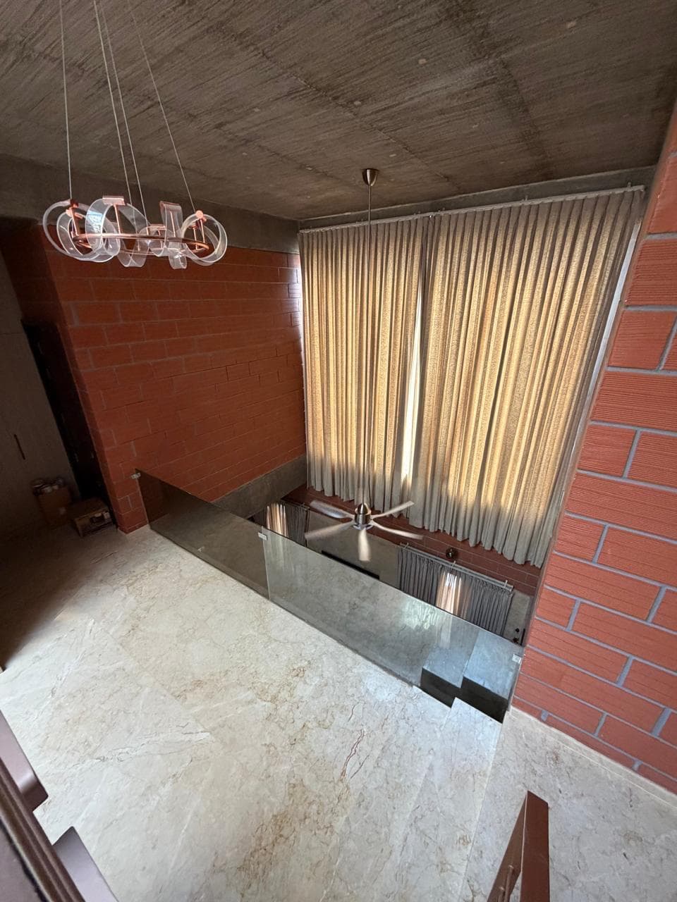 Double-height living space in the Thanjavur residence with exposed brick walls, sheer curtains, and a pendant light cluster.